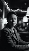 Professional businesswoman in elegant blazer sitting in upscale restaurant with bokeh lights creating atmospheric black and white portrait for corporate branding.