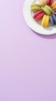 Colorful macarons arranged on white plate against soft lavender background. Assorted flavors in pastel shades including pistachio, raspberry, yellow, and blueberry. Overhead flat lay composition with ample copy space for bakery, confectionery, or dessert marketing.