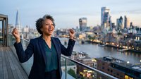 Woman with dark curly hair and medium skin tone wearing navy blazer and teal top celebrates success on rooftop terrace overlooking illuminated city skyline at dusk, arms raised in triumph and joy.