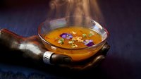 Steaming golden broth in a glass bowl with purple edible flowers, held in weathered hands, silver ring, dark moody background.