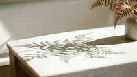 Young woman with medium skin tone, wearing casual attire, relaxing in minimalist bathroom with natural sunlight casting fern leaf shadows on white marble surface and potted plant.