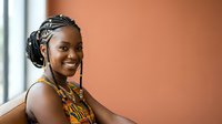 Young African woman with braided hairstyle and traditional beaded necklace smiling against warm orange background, suitable for diversity, culture, and lifestyle content.