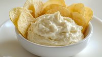 Creamy white dip served in ceramic bowl surrounded by crispy potato chips, close-up view on light background, appetizing party snack for entertaining.