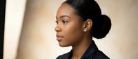 Young African American businesswoman in profile with professional hairstyle and dark blazer in modern office setting with warm natural lighting.