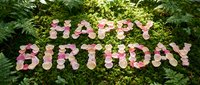 Happy birthday message spelled out in pink flowers arranged on green moss and foliage background for celebrations and greeting cards.