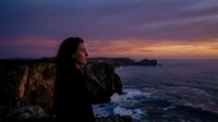 Man with long dark hair in profile gazing at dramatic purple and orange sunset over rocky coastal cliffs and ocean waves, contemplative mood for travel and nature themes.