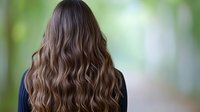 Young woman with long wavy brown hair viewed from behind in outdoor natural setting with soft bokeh background for hair care and beauty concepts.