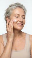 Mature woman with gray hair applying face cream with closed eyes and gentle smile, skincare routine for aging skin and anti-aging beauty treatment.