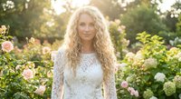 Young blonde Caucasian woman with curly hair in white lace dress standing in blooming rose garden during golden hour sunlight for romantic portrait photography.