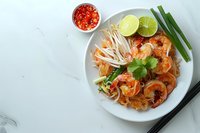 Traditional Pad Thai noodles with shrimp, bean sprouts, lime, and chili served on white plate against marble background, perfect for restaurant menu or food blog content.