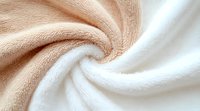 Soft beige and white twisted fabric creating spiral texture pattern. Close up view of plush microfiber material shows gentle folds and luxurious textile surface detail.