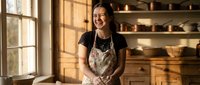Young Asian woman potter smiling in ceramic workshop wearing apron. Artisan craftsperson in pottery studio with shelves of clay bowls and natural lighting.