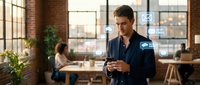 Young professional businessman using smartphone in modern office space with brick walls and large windows during golden hour lighting.