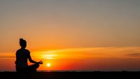 Woman silhouette meditating in lotus pose against vibrant orange sunset sky. Peaceful yoga practice outdoors for wellness and mindfulness concepts.