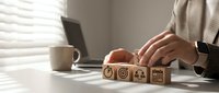 Businessperson arranging wooden cubes with business strategy icons on a desk, symbolizing goal setting, planning, and teamwork in a modern office.