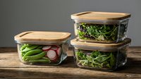 Glass food storage containers with bamboo lids containing fresh vegetables and greens for healthy meal prep and kitchen organization on wooden surface.