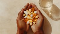 Person holding handful of vitamin supplements and capsules in various colors with glass of water on neutral background for healthcare and wellness concepts.