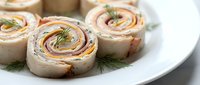 Sliced pinwheel wraps filled with cream cheese, ham, and fresh dill garnish arranged on a white plate for appetizers, catering, or lunch presentations.