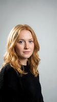 Professional portrait of confident young Caucasian woman with wavy blonde hair wearing black sweater against neutral gray background for business use.