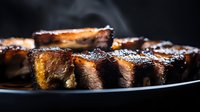 Juicy grilled pork ribs with caramelized BBQ sauce and charred edges on dark plate, close-up view showing tender meat texture and glossy glaze finish.