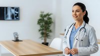 Female doctor in white coat with stethoscope standing in modern medical office with wooden desk and plant background for healthcare concepts.