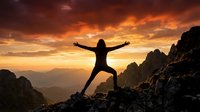 Silhouette of person with arms raised celebrating success on mountain peak at dramatic sunset with orange sky and rocky landscape for motivation concepts.
