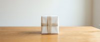 Small white gift box tied with a beige ribbon sitting on a wooden table against a light background, minimalist holiday present concept.