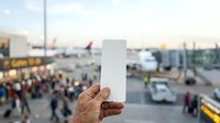 Mature male hand holding blank white card at airport terminal with planes, vehicles, and passengers in blurred background for travel, business, or advertising mockup purposes.