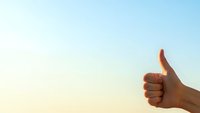 Hand showing thumbs up gesture against bright blue sky with warm sunlight creating positive approval sign for success and achievement concepts.