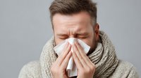 Caucasian man with closed eyes blowing nose into tissue, wearing beige scarf against gray background, showing symptoms of cold or seasonal allergies.