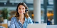 Young woman with headset smiling while working on laptop in modern office space. Customer service representative in professional environment.