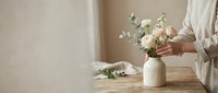 Caucasian woman arranging white ranunculus and eucalyptus in a ceramic vase on a rustic wooden table with neutral linen tones.