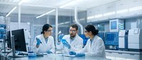 Three diverse medical researchers in white lab coats collaborate in modern laboratory, examining samples and data on computer screens for scientific discovery.