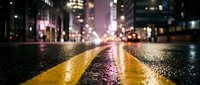 Wet city street at night with glowing yellow road markings, bokeh lights, and urban skyline in the background after rain.