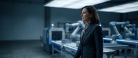 Female engineer or technician with long dark hair, wearing professional black blazer, standing in modern industrial facility with robotic arms and manufacturing equipment in background, representing automation, technology, and industry.