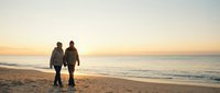 Couple walking hand in hand on sandy beach at golden sunset with ocean waves, romantic evening stroll by seaside creating peaceful silhouette against warm sky.