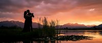 Romantic couple silhouette embracing at sunset by mountain lake with dramatic orange pink sky reflecting on calm water creating peaceful moment.
