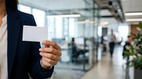 Young professional woman in dark blazer holding blank business card in modern office lobby with blurred colleagues and glass partitions in background.