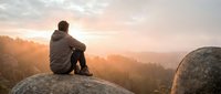 Man sitting on rock overlooking misty forest valley at sunrise, contemplating nature landscape with golden light filtering through trees.