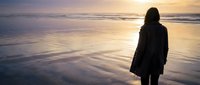 Woman standing alone on beach at sunset, silhouetted against golden sky and calm ocean waves, contemplative mood for wellness and lifestyle content.