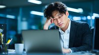 Young Asian businessman showing signs of stress and fatigue while working late at office desk with laptop computer in corporate environment.