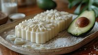 Handmade organic soap bars with floral pattern on wooden plate, fresh avocado and sea salt. Natural skincare and spa composition in warm light.