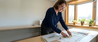Professional woman architect reviewing construction blueprints at wooden desk near window in modern office workspace for project planning.