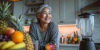 Smiling mature Asian woman with gray hair in modern kitchen surrounded by fresh tropical fruits and healthy ingredients for nutritious meal preparation.