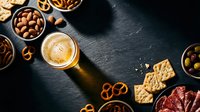 Beer glass with foam surrounded by assorted snacks including pretzels, nuts, crackers and olives on dark slate background for appetizer presentations.