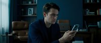 Young man of Caucasian ethnicity in casual dark blue shirt using smartphone in modern home office with blue walls, bookshelves and leather furniture for communication and technology concepts.