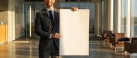 Professional businessman in dark suit holding blank white poster in modern office lobby with natural lighting for corporate presentations and marketing.