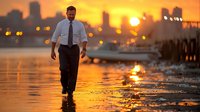 Caucasian businessman walking alone on wet pier at sunset, city skyline silhouette in background, orange sky reflects in water creating dramatic atmosphere.
