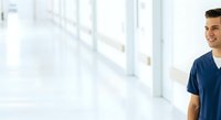 Young male healthcare professional, Caucasian ethnicity, wearing navy scrubs, smiling in bright modern medical facility corridor with large windows and natural lighting.