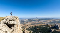 Lone hiker stands triumphantly on rocky mountain cliff edge overlooking vast wilderness landscape with rolling hills, forests and distant peaks against clear blue sky.